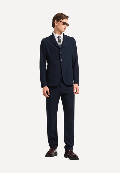 Giovane uomo che indossa un completo blu navy scuro, camicia bianca, cravatta con motivo, occhiali da sole e scarpe marroni scure, in piedi con le mani rilassate.
