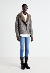 Grå syntetläderjacka med en brun fårskinnskrage, dragkedja fram och två sidofickor, bärs över en vit skjorta och blå jeans. Svarta ankelstövlar.