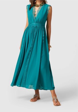 Femme portant une robe maxi sans manches couleur sarcelle avec finition en dentelle, collier chaîne en or et bracelet, et sandales plates ornées de bijoux sur fond uni.