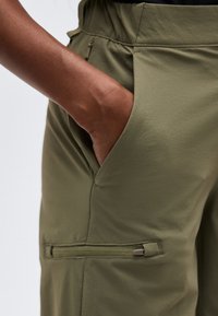 Χέρι με σκούρο χρώμα δέρματος που ακουμπά στην τσέπη παντελονιού ελιάς με φερμουάρ κάτω από την κύρια τσέπη.