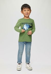 Grünes Langarm-T-Shirt mit dem Aufdruck "FRIENDSAURS" und blauen Pailletten; kombiniert mit hellblauen Jeans und weißen Sneakern.