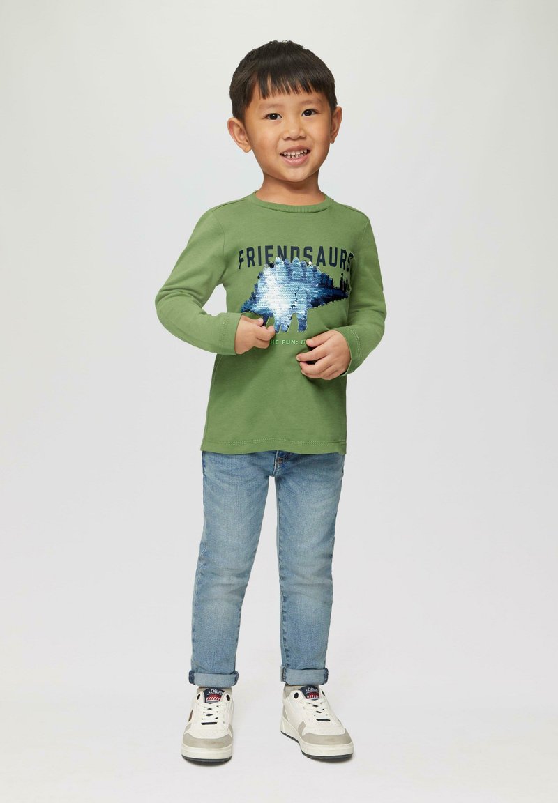 Groene longsleeve t-shirt met de tekst "FRIENDSAURS" en een blauwe paillettenontwerp; gecombineerd met lichtblauwe jeans en witte sneakers.