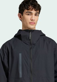 Jachetă neagră cu glugă, cu o textură netedă, având o închidere cu fermoar și un accent reflective pe partea stângă.