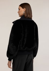 Bomber jacket in finta pelliccia nera con colletto e polsini a costine blu navy; presenta una vestibilità rilassata e una finitura morbida e testurizzata.