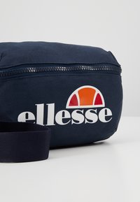 Sac banane en tissu bleu marine avec fermeture éclair noire, sangle ajustable et logo "ellesse" blanc avec un demi-cercle orange et rouge.