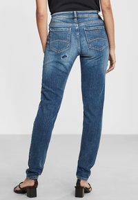 Modré džíny z denimu s úzkým střihem, vysokým pasem, zadními kapsami a malou poškozenou částí na pravé nohavici.