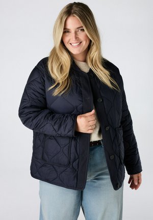 Femme souriante aux longs cheveux blonds portant une veste matelassée noire sur un pull beige et un jean bleu clair devant un fond uni.