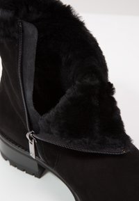 Bottine en daim noir avec doublure en fourrure noire douce, fermeture éclair argentée partiellement ouverte, et petit talon large sur fond blanc.