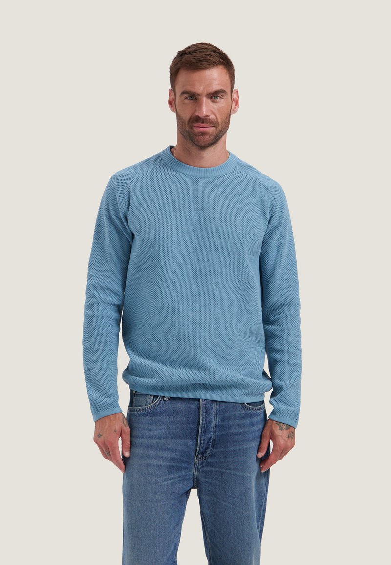 Homme portant un pull texturé bleu clair à manches longues et un jean bleu, debout devant un fond uni clair.