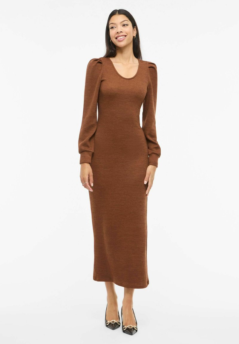 Ruskea, pitkähihainen neulemekko, jossa on pyöreä pääntie ja liehuvat olkapäät. Vartalonmyötäinen siluetti ja keskipitkä helma, teksturoitu kangas, yhdistetty mustiin kenkiin.