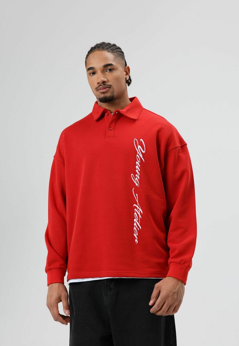 Rode sweatshirt met een polokraag, met een witte "Young Atelier" print langs de zijkant. De stof lijkt zacht; het ontwerp heeft een ontspannen pasvorm.
