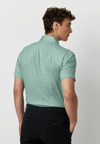 Korte mouwen shirt in lichtgroene stof met een gestructureerde afwerking, met een klassieke kraag en een rechte achterkant. Draagt met donkere broek.