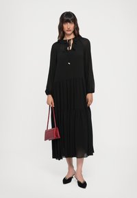 Vestito nero a maniche lunghe con scollatura arricciata, design a strati svasati e tessuto leggero. Abbinato a una piccola borsa rossa e scarpe nere.
