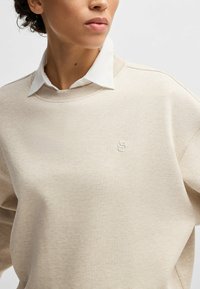 Felpa beige con superficie testurizzata, collo rotondo e rifiniture a coste. Presenta un piccolo logo ricamato bianco sul petto.