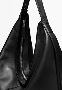Sac à main en cuir noir avec une bandoulière unique et un détail de fermeture éclair argentée visible sur la poche latérale.