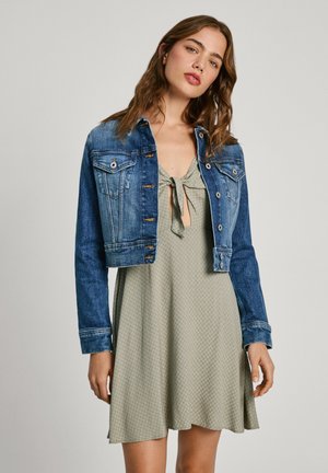 Cropped denim jacket in dark blue with front pockets and button closure, layered over a light green dress with a tied neckline and textured pattern.