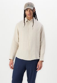 Beige linne skjorta med knappar och långa ärmar, lättviktsmaterial och rak fåll, i kombination med marinblå byxor. Har en subtil textur.
