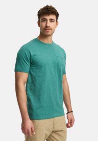 T-shirt in cotone turchese con collo a giro e maniche corte, dotato di taschino sul petto e una texture sottile. Abbinato a pantaloni beige.