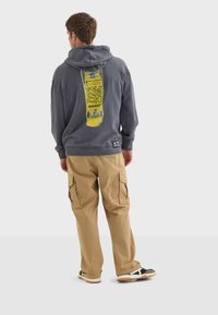 Felpa grigia con una grafica gialla di uno skateboard sulla schiena, abbinata a pantaloni cargo beige e sneakers nere con accenti bianchi.