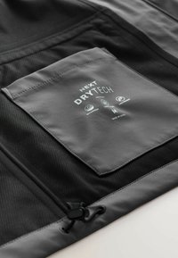 Gray jacket featuring a mesh texture with a pocket displaying "Next DryTech" branding, waterproof symbols, and pull toggle.
