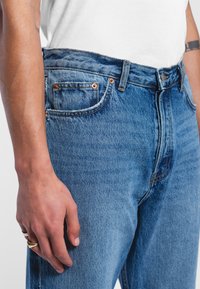 High-waisted blue denim jeans with copper rivet accents, a side pocket, and a faded texture. The fabric appears durable and thick.