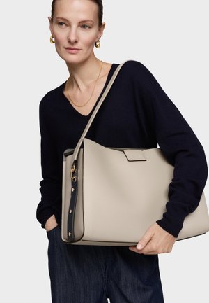 Mujer con pendientes de aro dorados y collar, sosteniendo un bolso grande beige con asa negra, vestida con suéter azul marino y vaqueros oscuros.