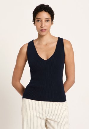 Frau mit kurzen lockigen Haaren, die ein marineblaues, geripptes, ärmelloses Oberteil und weiße Hosen mit dünnen vertikalen Streifen trägt, steht mit den Händen hinter dem Rücken.