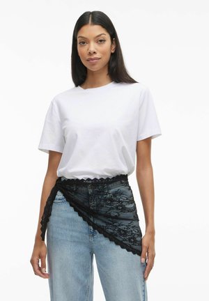 Sort blondebælte med bølgede kanter, båret over lyse denim jeans og en hvid t-shirt. Indviklede blomster mønstre synlige på blondestoffet.
