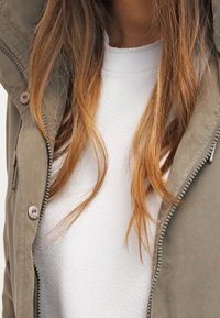 Khaki bunda se zipem a knoflíkovým uzávěrem, vybavená kapucí a strukturovanou tkaninou, nošená přes bílý top s hladkým povrchem. Jsou viditelné dlouhé hnědé vlasy.