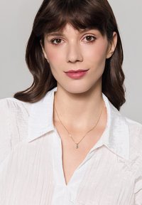 Złoty naszyjnik z małym turkusowym wisiorkiem, noszony na białej, fakturowanej koszuli z kołnierzykiem. Projekt jest delikatny i minimalistyczny.