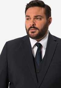 Dunkelblaues Sakko aus glattem Stoff. Mit Reverskragen, Ein-Knopf-Verschluss und einer Brusttasche. Kombiniert mit einem weißen Hemd und einer Krawatte.