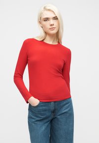 Νεαρός με ανοιχτόξανθα μαλλιά φοράει tight κόκκινο μακρυμάνικο πουκάμισο και μπλε τζιν, στέκεται με το ένα χέρι στην τσέπη.