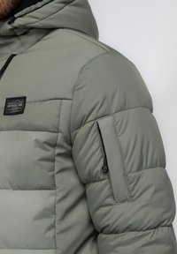 Chaqueta tipo puffer en gris verdoso con capucha, que presenta una gran textura acolchada, bolsillo lateral con cremallera y parche de marca en el pecho.