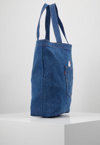 Sac fourre-tout en denim avec deux bretelles, une poche avant, couleur bleu clair, coutures visibles et petit détail de logo près du haut.