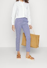 Bílá košile na knoflíky, modré vzorované kalhoty, hnědé sandály s tlustými podešvemi a pletená hnědá taška. Stojí na jednobarevném pozadí.