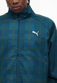 Νέος φορεί ένα μπλε και πράσινο καρό μπουφάν Puma με ψηλό γιακά, φερμουάρ μπροστά και λευκό λογότυπο Puma στο στήθος.