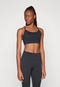 Mulher atlética vestindo um top desportivo preto e leggings de cintura alta, em pé e olhando para o lado contra um fundo simples.