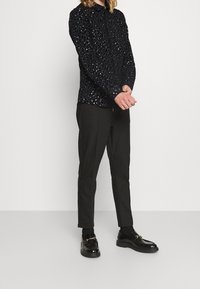 Černá dlouhá košile s texturovaným leopardím vzorem, spárovaná s úzkými černými kalhotami a lesklými černými mokasíny se zlatými akcenty.