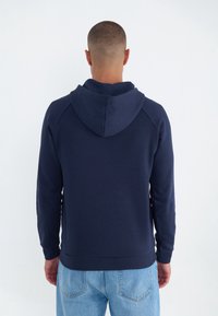 Man som vänder bort med ryggen iförd en marinblå luvtröja och ljusblå jeans mot en enkel vit bakgrund.