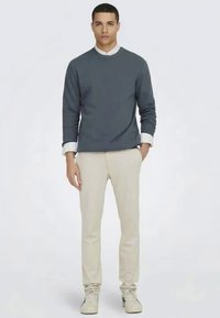 Mørkegrå crew-neck sweatshirt, hvid skjorte under, beige bukser og lyse sneakers, båret af en stående mandlig model.