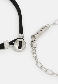 Bracelet en suede noir avec un fermoir en métal argenté et une chaîne argentée mettant en avant une étiquette rectangulaire gravée avec "ICON".