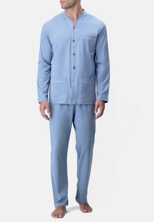 Uomo che indossa un pigiama azzurro chiaro a maniche lunghe con bottoni, tre tasche e pantaloni abbinati a tutta lunghezza, a piedi nudi su sfondo semplice.