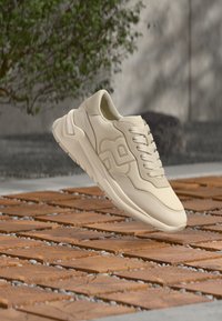 Baskets en cuir beige à texture lisse, bout rond, col matelassé et design à lacets. Dotées d'un logo et d'une semelle épaisse, posées sur une surface en bois.