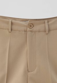 Primo piano di pantaloni beige con passanti per cintura e chiusura con bottone in vita.