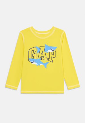 Gul langærmet børneskjorte med teksten "GAP" og to blå tegneseriehajer trykt på fronten.