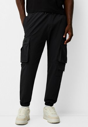 Mann trägt schwarze Cargo-Joggerhosen mit elastischen Bündchen und mehreren Taschen, kombiniert mit weißen Sneakers und einem schwarzen Oberteil.