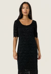 Musta pitsimekko, jossa on pyöreä pääntie ja lyhyet hihat, kukkakuviolla ja scallop-reunoilla. Lattiapituinen malli.