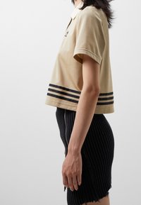 Zkrácená béžová polo tričko s límečkem a černými pruhy na spodním lemu, spárované s černou pletenou sukní s bočním zipem.