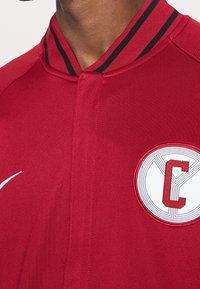 Rød jakke med opretstående krave, sort og hvide striber. Har et cirkulært mærke med et "C" logo på brystet. Glat stoftekstur.