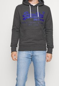 Felpa con cappuccio grigio scuro con lacci bianchi, grande testo/design blu sulla parte anteriore, tasca frontale, polsini e orlo a coste. Jeans in denim chiaro.